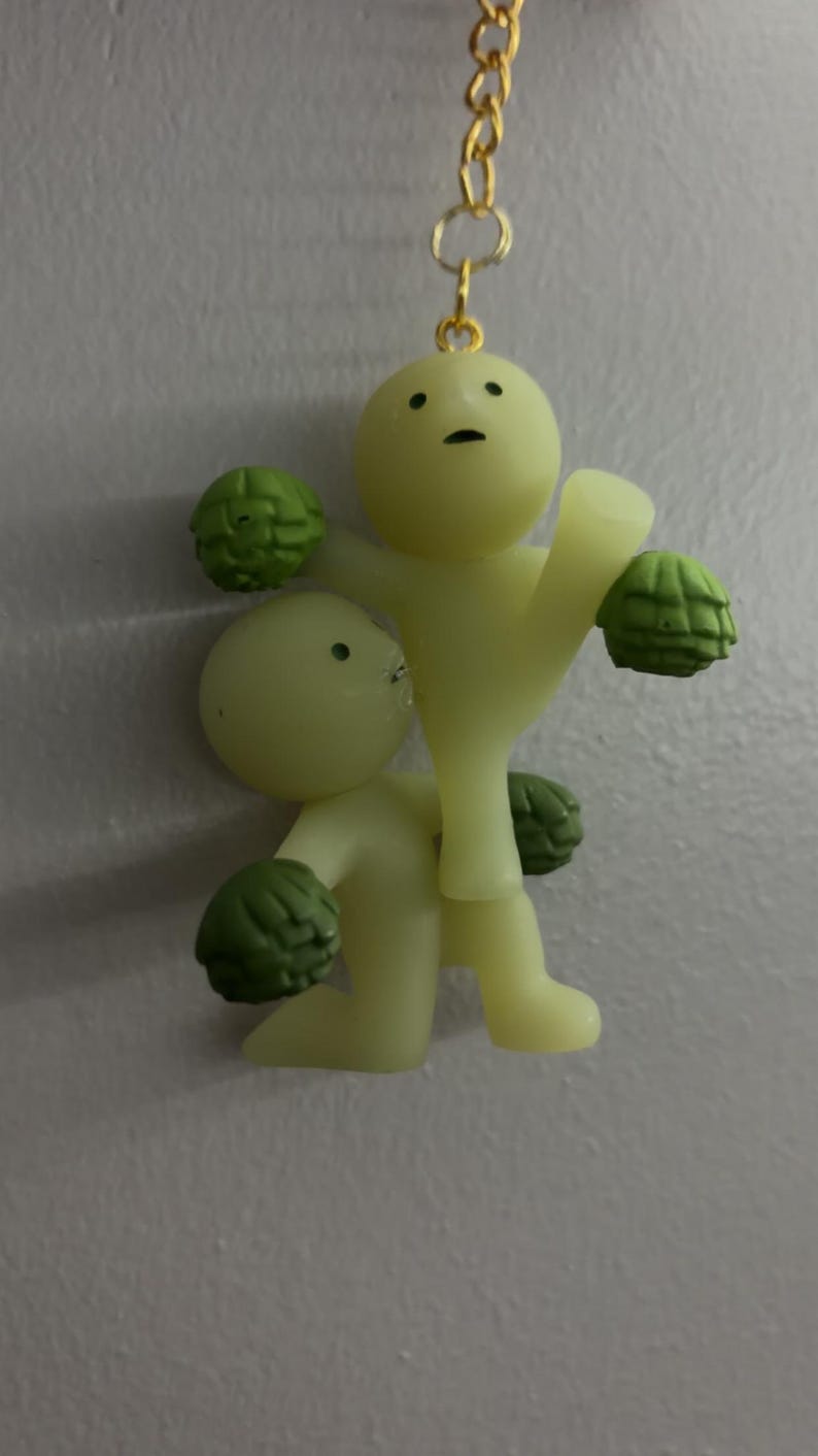 May include: A pale yellow keyring featuring two small, smooth figures with simple facial features. One figure stands while the other kneels, both holding textured green spherical objects resembling artichokes. A gold-toned metal chain is attached.