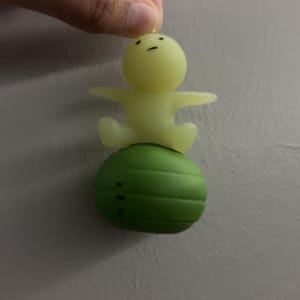May include: A small, pale yellow, translucent figurine with a simple face featuring two dots for eyes and a line for a mouth, sitting atop a solid, dark green, spherical object with horizontal ridges that resembles a small exercise ball.