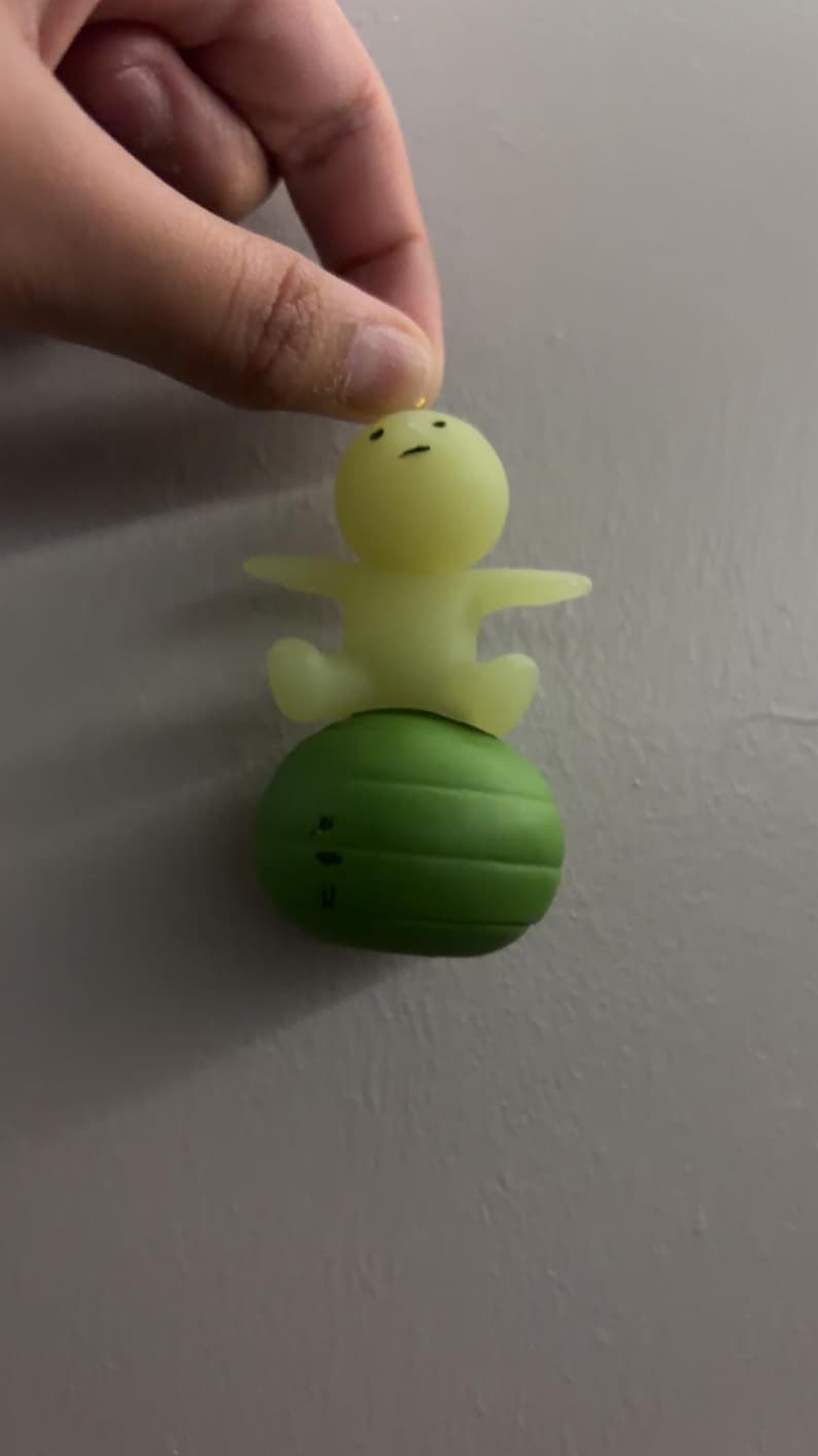 May include: A small, pale yellow, translucent figurine with a simple face featuring two dots for eyes and a line for a mouth, sitting atop a solid, dark green, spherical object with horizontal ridges that resembles a small exercise ball.