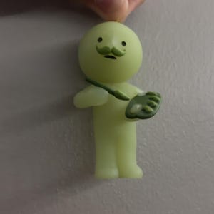 May include: A small, pale yellow, glow-in-the-dark figurine of a round-headed character with a green moustache. The character holds a green paintbrush in one hand and a green artist palette with white paint dabs in the other hand.