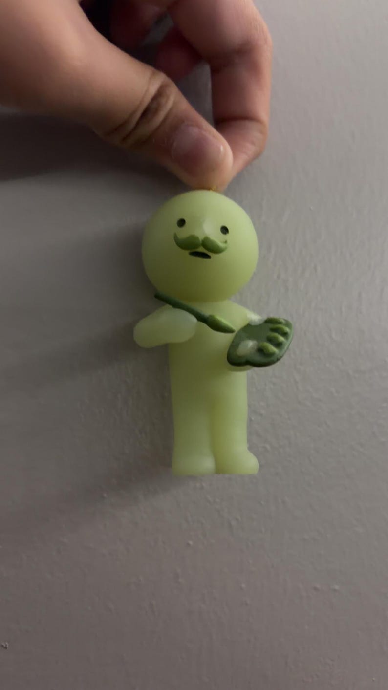 May include: A small, pale yellow, glow-in-the-dark figurine of a round-headed character with a green moustache. The character holds a green paintbrush in one hand and a green artist palette with white paint dabs in the other hand.