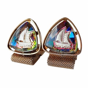 Può includere: Un paio di gemelli vintage a tema nautico. Ogni gemello presenta un inserto triangolare in vetro iridescente con un disegno di barca a vela argentata, incastonato in una montatura in metallo dorato. I gemelli sono attaccati a un retro in rete dorata.