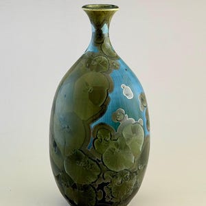 May include: A ceramic vase with a narrow neck and an oval body, featuring a crystalline glaze. The surface displays a vibrant blue background overlaid with large, circular, olive-green and dark brown crystalline patterns throughout the exterior.