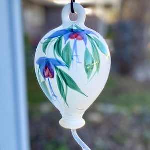 May include: White ceramic hummingbird feeder with hand-painted blue and red floral design. The feeder has a clear plastic tube with a red tip and a black hanging cord. A decorative garden accent.