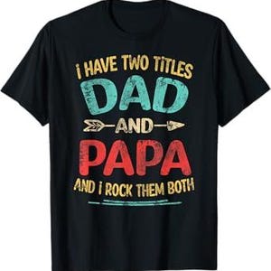 Pode incluir: Uma t-shirt preta de manga curta com o texto I HAVE TWO TITLES DAD AND PAPA AND I ROCK THEM BOTH em letras desgastadas amarelas, azul-petróleo e vermelhas, com um pequeno gráfico de seta separando as palavras DAD e PAPA na frente.