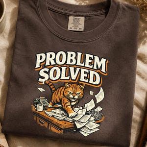 Könnte beinhalten: Ein dunkelgraues T-Shirt mit einer Grafik eines orangefarbenen getigerten Katers, der auf einem mit verstreuten Papieren bedeckten Schreibtisch steht. Über dem Kater steht der Text "PROBLEM SOLVED" in cremefarbenen Buchstaben mit orangefarbenem Rand.