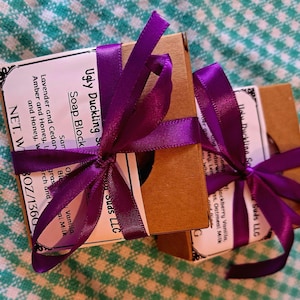 May include: Two square brown cardboard boxes containing soap blocks, each wrapped with a vibrant purple satin ribbon tied in a bow. The white labels feature the text Ugly Duckling Suds LLC and list ingredients, with a net weight of 4.8 ounces.