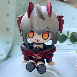 Può includere: Una piccola statuetta in stile chibi raffigurante un personaggio con capelli grigi corti, due protuberanze rosse a forma di corna e grandi occhi arancioni. La figura indossa un abito scuro con accenti rossi, un ampio colletto e stivali scuri.
