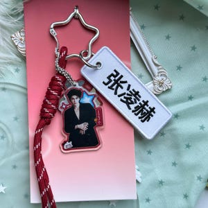 May include: A keychain featuring a star-shaped metal ring, a red braided cord, a clear acrylic charm with a portrait of a person in a black suit, and a white rectangular fabric tag embroidered with the black Chinese characters 张凌赫.