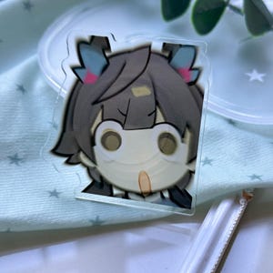 May include: A clear acrylic charm featuring a stylized anime-inspired character with dark gray hair, prominent light-colored eyes, and small blue and pink accents on the hair. The character has a neutral expression with a small, light-colored mouth.
