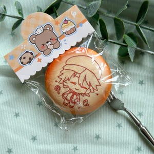 Puede incluir: Un objeto redondo de color naranja con forma de macarrón en una bolsa de plástico transparente con una etiqueta de cartón. La etiqueta muestra un oso de dibujos animados, un cupcake y una galleta. El macarrón tiene un dibujo lineal rojo de un personaje.