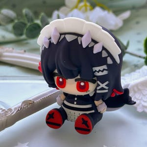 May include: A small, stylized figurine featuring a character with dark hair, bright red eyes, and a white headpiece. The figure wears a black and white outfit with red accents on the feet, and displays the Roman numeral XI on the side of the hair.