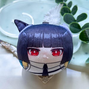 May include: A round plush toy featuring a character with short black hair, pointed cat-like ears, and bright red eyes. The character wears a white collared shirt with a black tie and blue accents. The plush has a soft, smooth texture and a spherical shape.