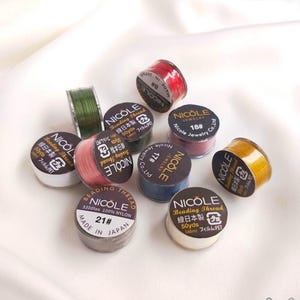 Puede incluir: Un conjunto de carretes de hilo para abalorios Nicole en varios colores, como rojo, verde y rosa. Cada carrete está etiquetado con el número de hilo y el texto "Beading Thread" en inglés y japonés. Fabricado en Japón.