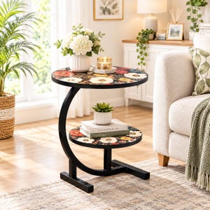 May include: A round two-tier side table with a black metal C-shaped frame and floral patterned surfaces featuring red, white, and orange flowers on a black background. The table holds a white vase with flowers, a candle, and two books.