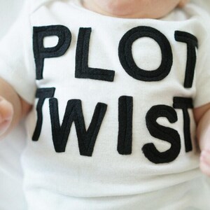 K&ouml;nnte beinhalten: Wei&szlig;er Baby-Body mit den Worten "PLOT TWIST" in gro&szlig;en, schwarzen Blockbuchstaben. Der Body hat kurze &Auml;rmel und das Baby liegt. Die H&auml;nde des Babys sind sichtbar.