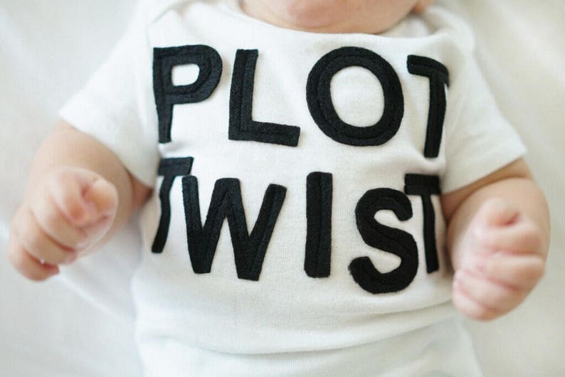 K&ouml;nnte beinhalten: Wei&szlig;er Baby-Body mit den Worten "PLOT TWIST" in gro&szlig;en, schwarzen Blockbuchstaben. Der Body hat kurze &Auml;rmel und das Baby liegt. Die H&auml;nde des Babys sind sichtbar.