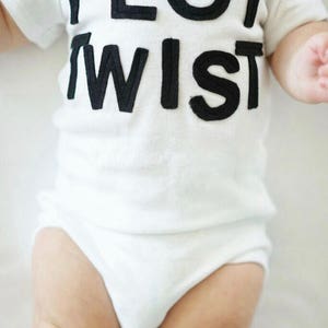 K&ouml;nnte beinhalten: Ein wei&szlig;er Baby-Body mit dem Schriftzug "PLOT TWIST" in gro&szlig;en, schwarzen Blockbuchstaben. Der kurz&auml;rmelige Body bedeckt den Oberk&ouml;rper und die Beine des Babys und bietet ein schlichtes, klares Design.