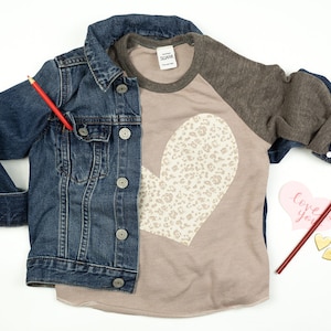 May include: A denim jacket and a raglan shirt with a heart design. The shirt has a taupe body, gray sleeves, and a heart-shaped leopard print. A red pencil and a pink heart-shaped card with "love you" are also present.