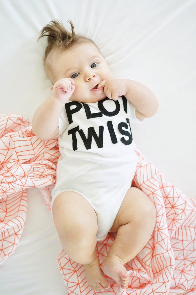 K&ouml;nnte beinhalten: Ein Baby in einem wei&szlig;en Body mit dem schwarzen Aufdruck "PLOT TWIST". Das Baby liegt auf einem wei&szlig;en Laken mit einer korallenfarbenen und wei&szlig;en geometrisch gemusterten Decke. Das Baby hat einen Haarschopf und lutscht an seinen Fingern.