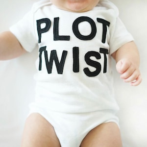 K&ouml;nnte beinhalten: Ein wei&szlig;er Baby-Body mit dem Text "PLOT TWIST" in gro&szlig;en, schwarzen Blockbuchstaben. Der kurz&auml;rmelige Body bedeckt den Oberk&ouml;rper und die Beine des Babys. Das Baby liegt auf einer wei&szlig;en Oberfl&auml;che.