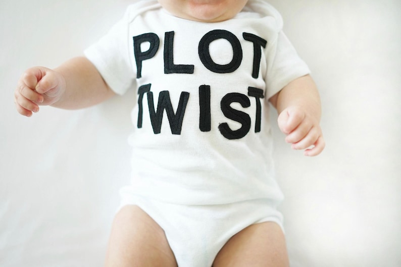 K&ouml;nnte beinhalten: Ein wei&szlig;er Baby-Body mit dem Text "PLOT TWIST" in gro&szlig;en, schwarzen Blockbuchstaben. Der kurz&auml;rmelige Body bedeckt den Oberk&ouml;rper und die Beine des Babys. Das Baby liegt auf einer wei&szlig;en Oberfl&auml;che.