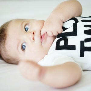 K&ouml;nnte beinhalten: Ein Baby mit blauen Augen tr&auml;gt ein wei&szlig;es T-Shirt mit den Worten "PLAY TIME" in gro&szlig;en schwarzen Buchstaben. Das Baby liegt auf einer wei&szlig;en Oberfl&auml;che, eine Hand in der N&auml;he des Mundes.