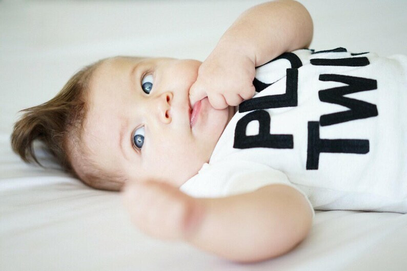 K&ouml;nnte beinhalten: Ein Baby mit blauen Augen tr&auml;gt ein wei&szlig;es T-Shirt mit den Worten "PLAY TIME" in gro&szlig;en schwarzen Buchstaben. Das Baby liegt auf einer wei&szlig;en Oberfl&auml;che, eine Hand in der N&auml;he des Mundes.