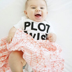 K&ouml;nnte beinhalten: Ein l&auml;chelndes Baby in einem wei&szlig;en Body mit den schwarzen Worten "PLOT TWIST". Das Baby ist in eine wei&szlig;-orange gemusterte Decke gewickelt. Das Baby hat einen B&uuml;schel braunes Haar.