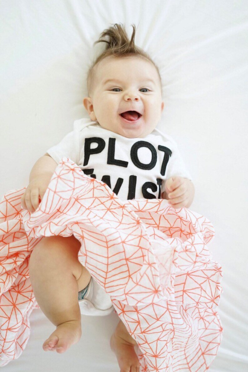 K&ouml;nnte beinhalten: Ein l&auml;chelndes Baby in einem wei&szlig;en Body mit den schwarzen Worten "PLOT TWIST". Das Baby ist in eine wei&szlig;-orange gemusterte Decke gewickelt. Das Baby hat einen B&uuml;schel braunes Haar.