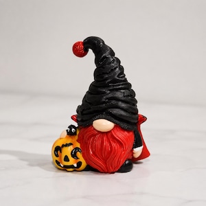 May include: A vampire-themed gnome figurine featuring a tall, textured black hat with a red glitter pom-pom, a long red beard, and a red cape. The gnome stands next to a small orange jack-o'-lantern pumpkin with a black spider on top.