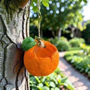 May include: An orange-coloured bird feeder, shaped like an orange, hanging from a tree. It has a textured surface, a green leaf, and a natural fibre string. The background shows a garden with a pathway.