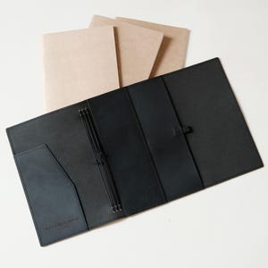 Puede incluir: Un diario de cuero negro abierto, que revela bolsillos y presillas elásticas para bolígrafos. Tres cuadernos de color canela son visibles detrás del diario. El diario tiene la palabra "RETENTEMPS" impresa en la parte delantera. El diario está sobre una superficie blanca.