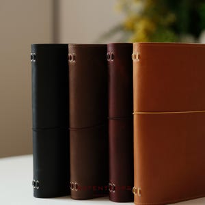 Könnte beinhalten: Vier Lederjournale in Schwarz, Braun, Burgunderrot und Hellbraun. Jedes Journal hat einen genähten Rücken und einen Verschluss. Die Journale stehen auf einer weißen Oberfläche mit unscharfem Hintergrund.