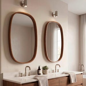 May include: A floating double vanity cabinet made of dark wood with a white countertop, two integrated sinks, and two silver taps. Above the vanity are two oval mirrors with dark wood frames and two wall-mounted silver light fixtures.