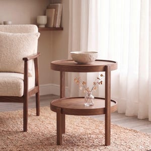 May include: A round, two-tiered wooden side table with a dark brown finish. The table features a circular top surface and a matching lower shelf supported by four straight legs. A textured off-white bowl sits on the top tier, and a small glass vase with dried stems rests on the lower shelf.