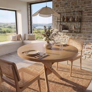 May include: A round light wood dining table with a cross-base design, accompanied by two chairs featuring woven cane backrests and seats. The table is set with ceramic plates, clear glasses, silverware, and a central vase holding dried flowers.