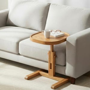 May include: A light grey two-seater sofa with a wooden side table. The table has a round top with a cup, glasses, and a book. A potted plant is in the background. The sofa is on a light-coloured rug.