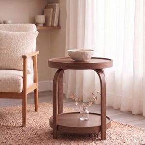 May include: A round wooden side table with a dark brown finish, featuring a raised edge on the top surface, a lower shelf, and four curved legs. A white textured bowl sits on the top, and a small clear glass vase with dried stems sits below.