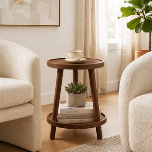 Peut inclure: Une table d'appoint ronde en bois foncé avec trois pieds et deux niveaux. Le plateau supérieur porte une tasse et une soucoupe en céramique crème. Le niveau inférieur contient une petite plante succulente verte dans un pot gris et trois livres empilés.