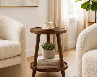 May include: A round dark wood side table with three legs and two tiers. The top tier holds a cream-colored ceramic cup and saucer. The bottom tier holds a small green succulent in a gray pot and three stacked books titled Kinfolk, Live Beautiful, and This Is Home.