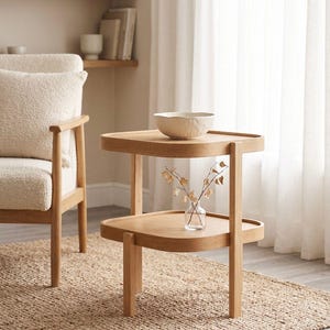 May include: A light oak side table with two square tiers featuring rounded corners and raised edges. The table has four straight legs and holds a textured off-white bowl on the top tier and a small clear glass vase with dried stems on the bottom.