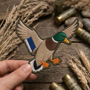 May include: An embroidered patch featuring a mallard duck in flight with wings spread. The duck has a green head, a yellow beak, a brown chest, tan body feathers, a blue wing patch, and orange webbed feet. The patch has a dark green border.