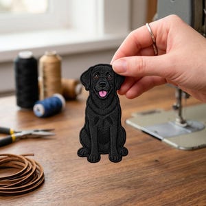 May include: An embroidered patch featuring a black Labrador retriever sitting in a forward-facing position. The dog has brown eyes, a black nose, and a pink tongue visible in its open mouth. The embroidery uses black thread for the coat.