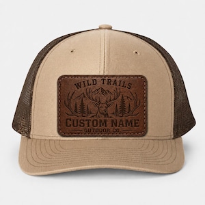 May include: A tan and brown trucker hat with a mesh back. The front features a brown leather patch with the words "WILD TRAILS" and "CUSTOM NAME" above a deer illustration. The hat is a classic, outdoor-themed accessory.