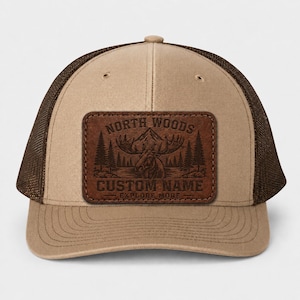 May include: A tan and brown trucker hat with a brown mesh back. The front features a rectangular leather patch with the words "NORTH WOODS" and "CUSTOM NAME" above and below a mountain and moose graphic. The text "EXPLORE MORE" is at the bottom.
