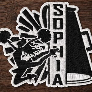 May include: An embroidered patch featuring a black and white design of a cheerleader holding pom-poms and jumping next to a megaphone. The megaphone has the name SOPHIA written vertically in bold black capital letters on a white background.