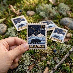 May include: A rectangular embroidered patch featuring a dark blue night sky with a yellow star constellation, white snow-capped mountains, and a black bear silhouette. The text reads 25 USA and ALASKA in yellow, with a scalloped white border.