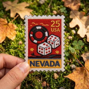 May include: A rectangular embroidered patch shaped like a postage stamp featuring a red background with a black and white poker chip, two white dice with red dots, and the text 25 USA. The bottom yellow section displays the word NEVADA in blue.