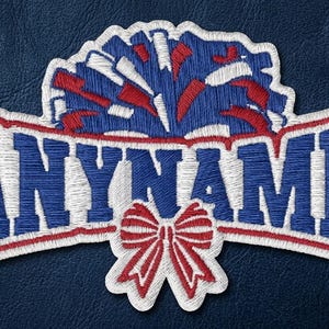 May include: An embroidered patch featuring the text ANYNAME in bold blue letters with a white outline. Above the text is a blue and red pom-pom, and below the text is a red and white bow. The entire design is bordered by a white stitched edge.